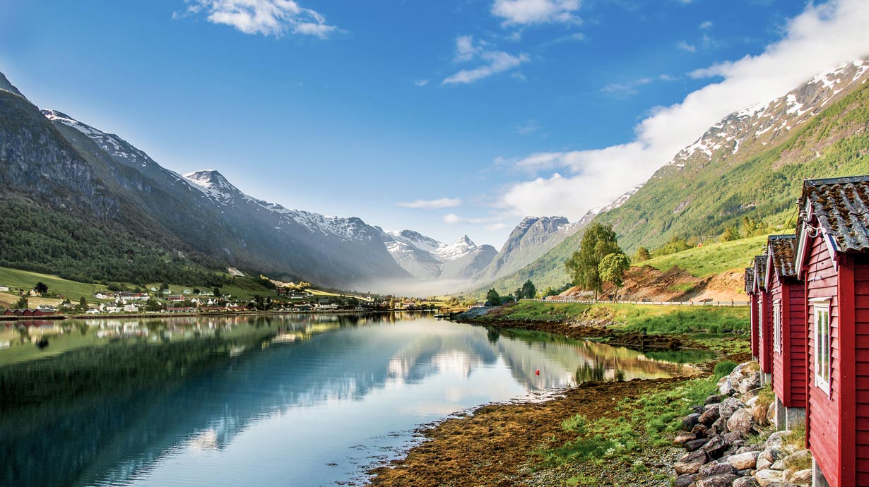 Scenic fjord with red cabins, snow-capped mountains, and calm water reflections | MSC Cruises Scenic fjord with red cabins, snow-capped mountains, and calm water reflections | MSC Cruises